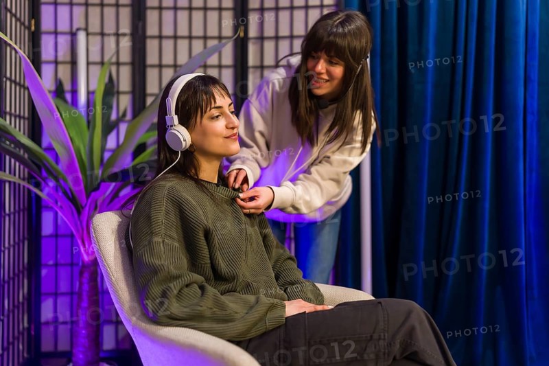 Young women setting up audio equipment, including headphones and a lavalier microphone, for a podcast or social media content session in a modern studio with purple lighting