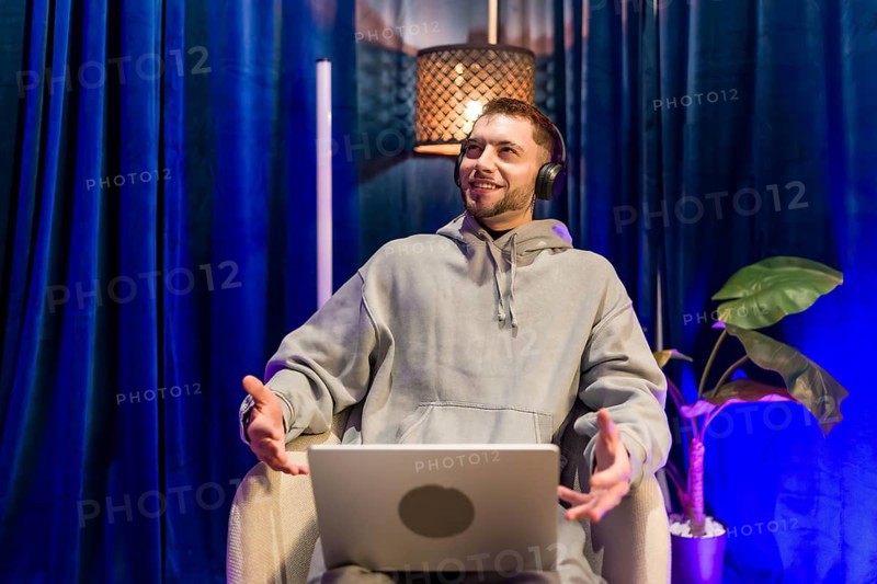 Young man wearing headphones and hoodie sitting with open laptop, actively speaking and gesturing during a live session or content creation in a modern studio