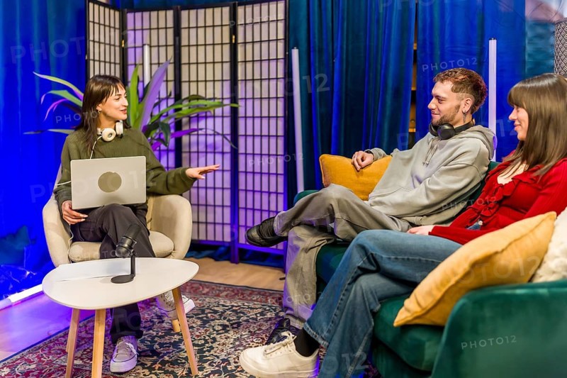 Young diverse creators recording a podcast in a stylish studio, chatting around microphones and laptops, collaborating on digital content for streaming and social media audiences