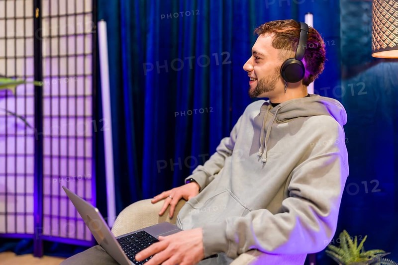 Young man creating digital content, recording a podcast or live stream, smiling and using a laptop while wearing headphones in a modern studio setting with blue and purple lighting