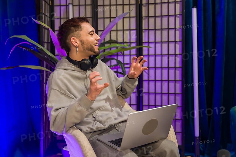 Young man wearing headphones and using a laptop, actively gesturing while recording a podcast or live streaming social media content in a modern studio