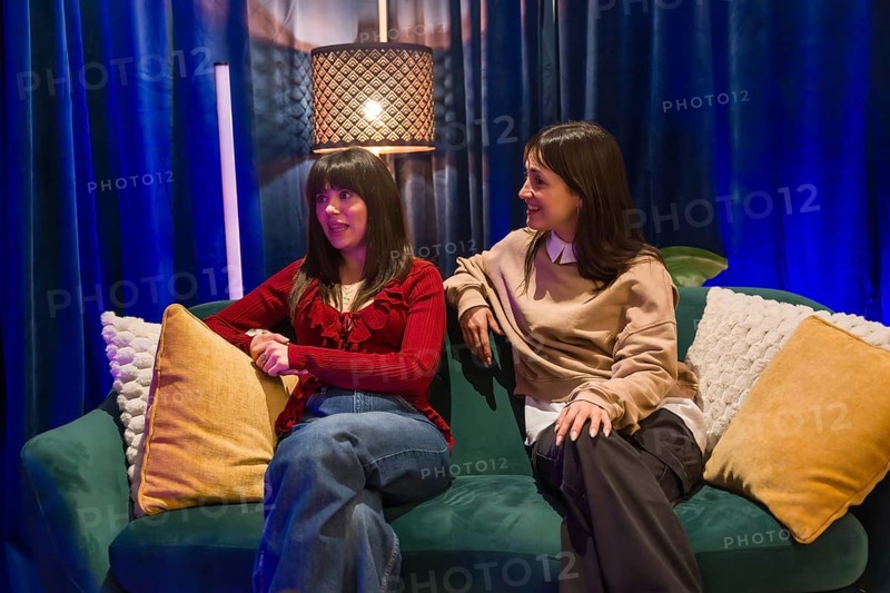 Two women content creators sitting on a green couch in a modern studio, engaging in conversation for a social media video or podcast recording, creating engaging digital content