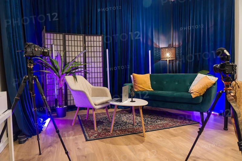 Modern content creation studio equipped for podcast recording or social media video production, featuring cameras on tripods, a microphone, laptop, sofa, armchair, and blue velvet curtains
