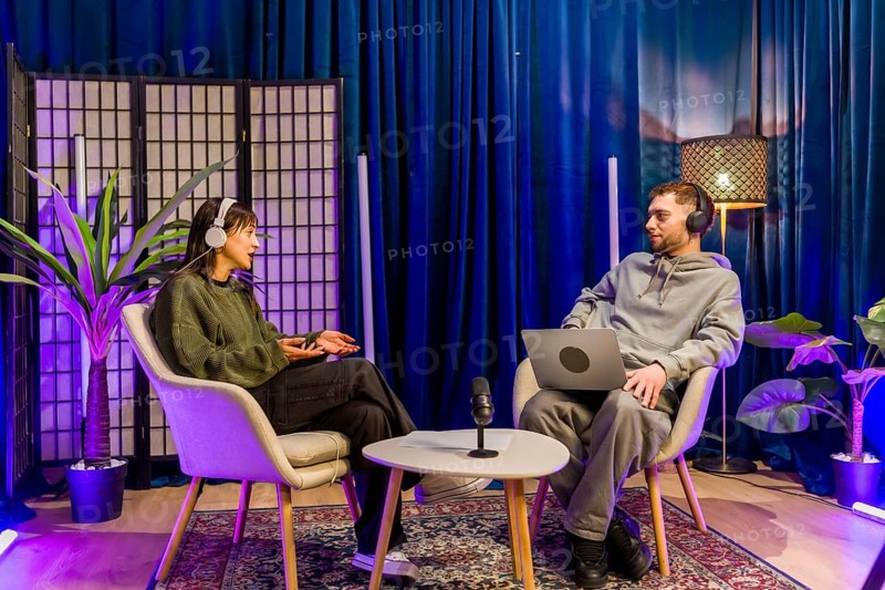 Two young adult content creators, a man and a woman, wearing headphones and sitting in chairs, are recording an audio podcast interview in a modern studio environment with a microphone and laptop