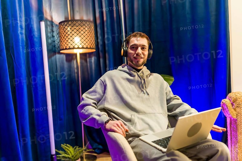 Young man wearing headphones and hoodie, sitting comfortably with a laptop in a home studio, smiling while streaming or recording a podcast for social media content