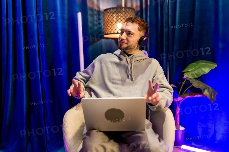 Young man wearing headphones and using laptop, recording a podcast or live streaming content from his creative home studio with colorful ambient lighting