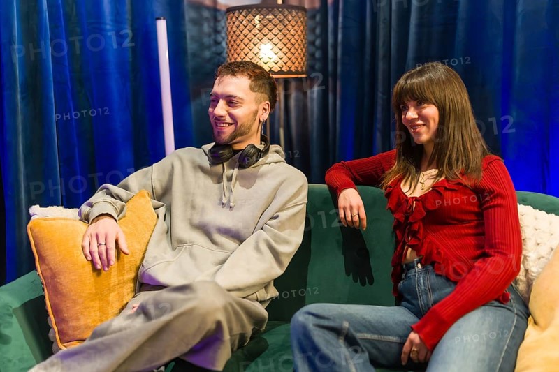 Young man and woman sitting on a green couch, sharing ideas and smiling during a social media content creation or podcast recording session in a modern studio