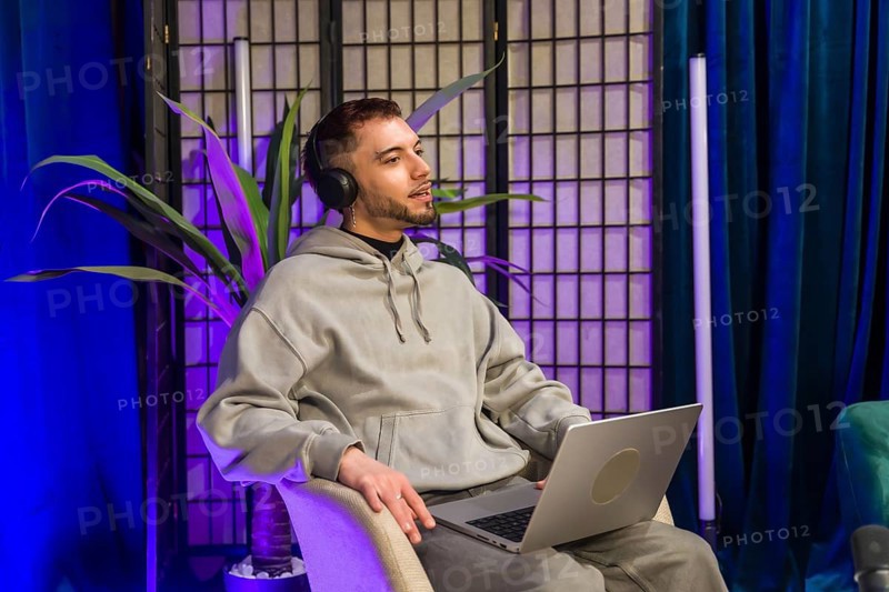 Young man wearing headphones and holding a laptop, talking and recording content for a podcast or social media, illuminated by blue and purple lights in a modern studio setup