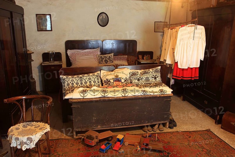 Traditional Transylvanian-Saxon farmhouse. The central bed is richly furnished with ornate pillows and blankets. There is a large, dark wooden chest in front of the bed. Such chests were historically used to store a bride's tax, clothing or valuables and were often hand painted or carved, museum within the fortified church of Honigberg, Biserica Fortificata Harman, Transylvania, Romania