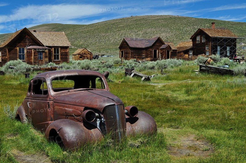 Rusty car wreck, Chevrolet Coupé, on green field in abandoned town with wooden houses, Bodie Ghost Town, California, USA