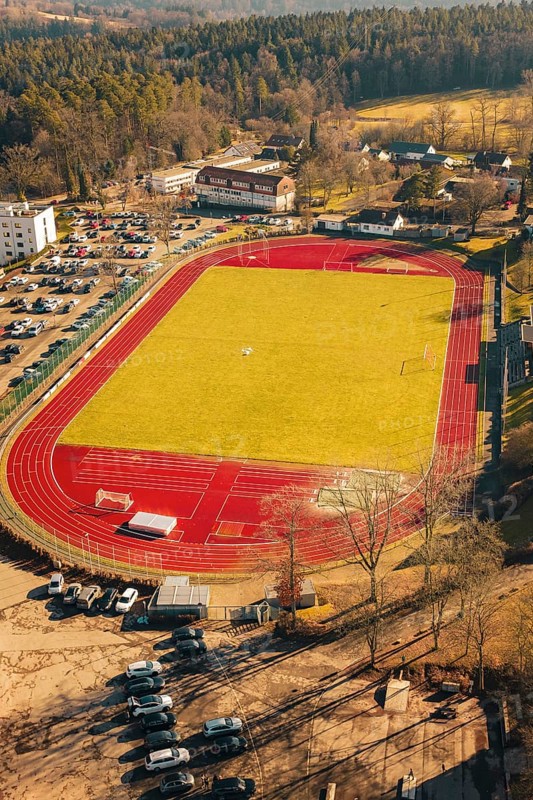 View of a large sports field with green field and red track, surrounded by cars and nature in winter, Wimberg, Calw, Germany