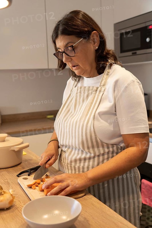 Vertical portrait of mature woman wearing apron while cutting carrots and potatoes in modern kitchen. Healthy homemade food, everyday lifestyle and copy space