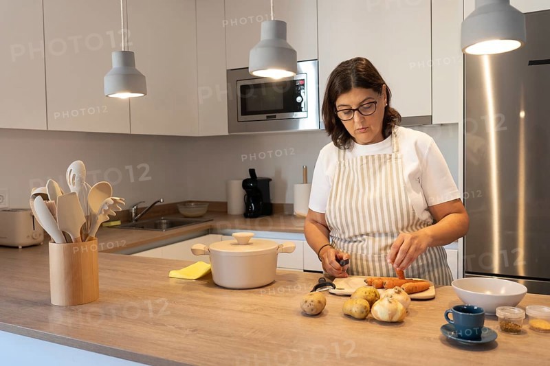 Mature woman wearing apron cutting carrots and potatoes while cooking at home in a modern kitchen. Everyday lifestyle, healthy food preparation, real domestic scene with warm light and copy space