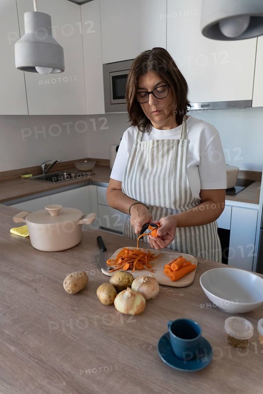 Woman wearing apron preparing vegetables for homemade soup while peeling carrots in modern kitchen. Simple ingredients and healthy home cooking concept