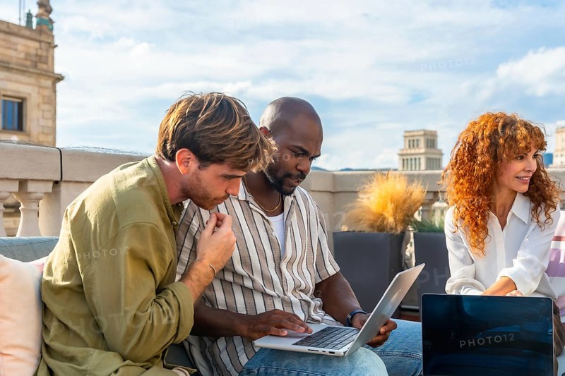 Diverse colleagues gathered on a rooftop coworking space focus on a laptop, collaborating on a project and brainstorming strategy in a casual, modern outdoor office setting