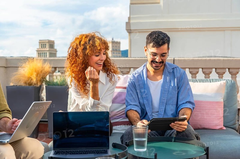 Young business colleagues working together on laptops and tablets, sharing ideas, and enjoying a casual outdoor meeting during their break on a modern urban rooftop coworking space