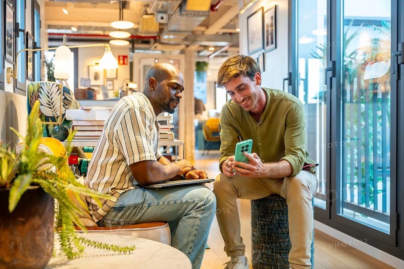 Two diverse male colleagues are sharing information on a mobile phone and laptop while working together, fostering teamwork and professional communication in a modern coworking space