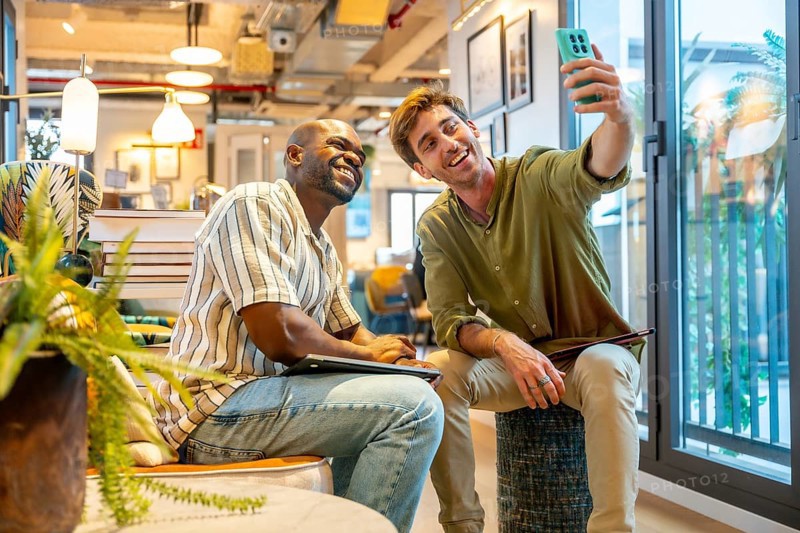 Two smiling multiracial men, professional colleagues, taking a selfie together with a smartphone while sitting in a contemporary coworking office space, representing teamwork and friendship