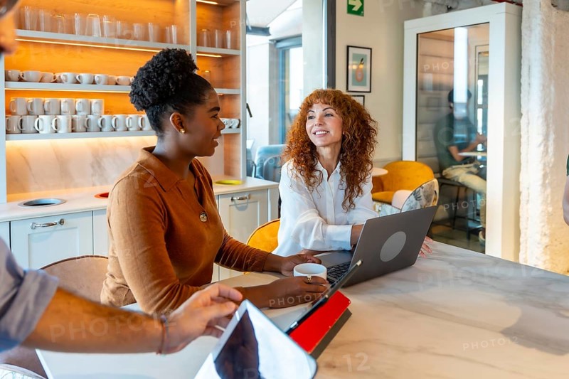 Multiracial businesswomen colleagues collaborating and discussing work around a laptop in a modern coworking kitchen while enjoying a coffee break, symbolizing teamwork and office life