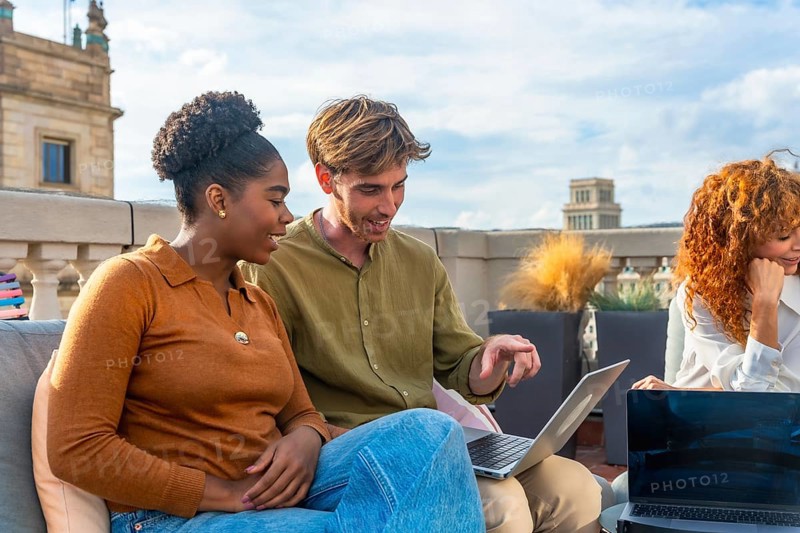 Diverse colleagues collaborate on a laptop during a rooftop meeting, sharing ideas and planning in a relaxed, sunny urban setting that blends work and social connection