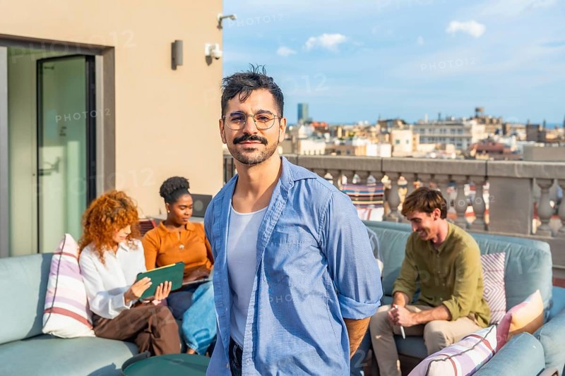 Smiling man standing on a rooftop terrace as diverse colleagues relax and work in the background, representing modern coworking, teamwork, and a pleasant work environment