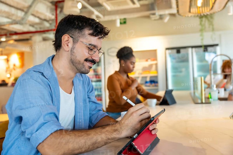 Man in casual attire smiling while using a digital tablet with a stylus to sketch ideas at a bar height table in a modern coworking space during a collaborative break