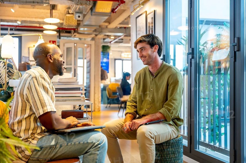 Two diverse male colleagues collaborate and chat during an informal break in a modern coworking office, sharing ideas, smiling and planning projects in a relaxed, professional setting