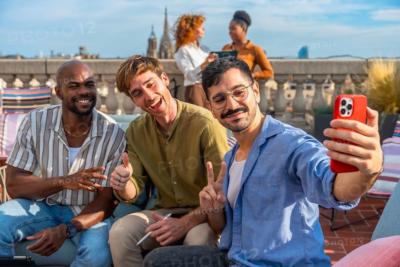 Diverse team of colleagues smiling and taking a rooftop selfie on a sunny urban terrace during a relaxed work break, enjoying casual conversation and city views