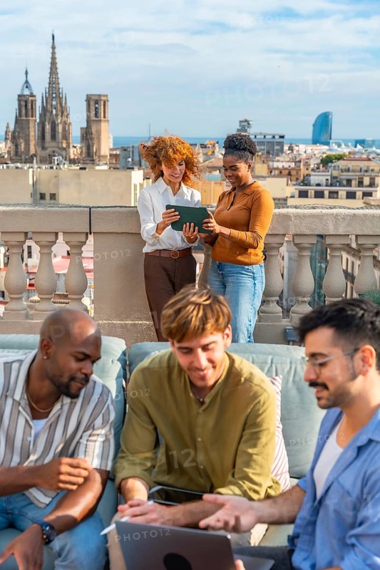 Diverse colleagues are brainstorming and working together with a laptop and tablet on a rooftop, enjoying a break with a city skyline view in the background