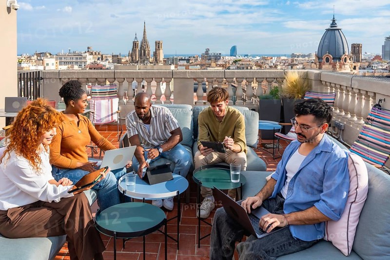 Diverse colleagues brainstorm and collaborate on laptops and tablets on a vibrant rooftop terrace in barcelona, enjoying a sunny break, city views and casual teamwork spirit