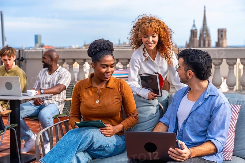 Diverse business professionals collaborating and networking on a barcelona rooftop terrace, using laptops and tablets for a casual outdoor meeting that blends teamwork and city lifestyle