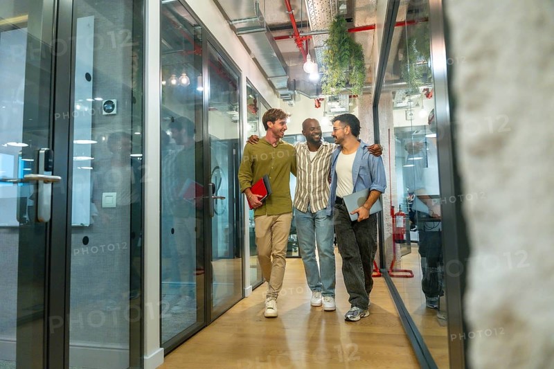 Three smiling diverse colleagues walking together embraced through a modern office building hallway, sharing a moment of friendship and teamwork during their work break