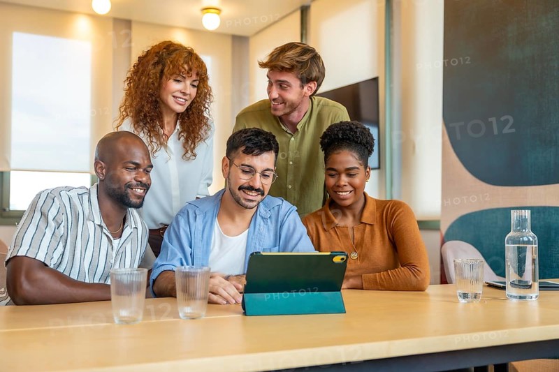 Diverse business team members gathering around a digital tablet, smiling and collaborating while discussing work during an office meeting or casual break