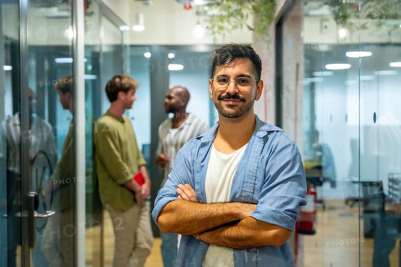 Confident male professional standing with arms crossed, smiling at the camera while his multi ethnic colleagues are talking and networking in the modern coworking office space