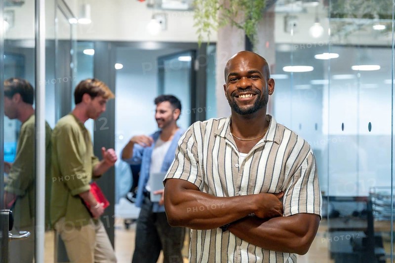 Smiling black businessman posing with arms crossed while diverse casual colleagues are communicating in the background of a modern glass office or coworking space during a break