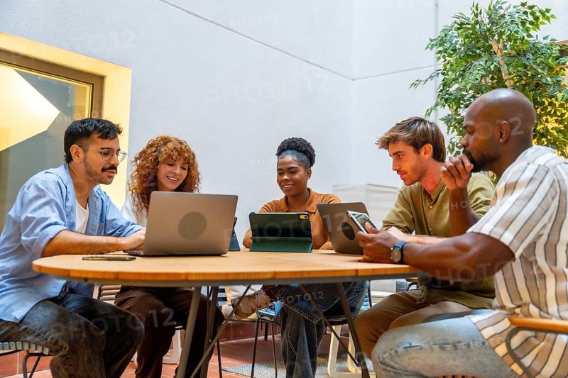 Diverse business colleagues working together on laptops and tablets at a round table, sharing ideas and collaborating during an informal meeting or break at their modern office or coworking space