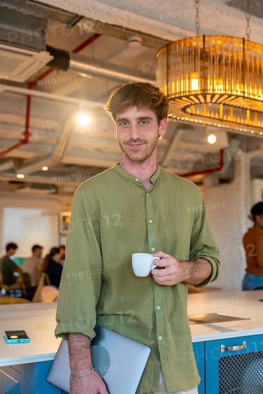 Young man standing in a modern coworking office, enjoying a coffee break while holding a laptop, surrounded by colleagues creating a dynamic work environment during a collaborative meeting