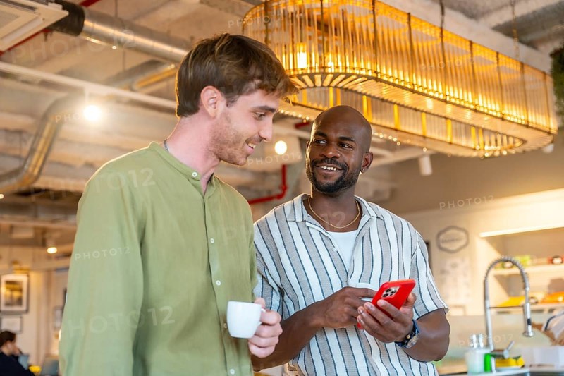 Two diverse male coworkers smiling and engaging in conversation while one holds a smartphone and the other holds a coffee cup in a modern office or coworking space during a casual break