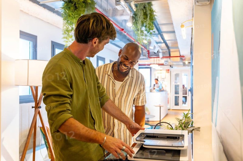 Male coworkers collaborating on a document at the printer, sharing a laugh and enjoying positive interaction in a modern coworking space, illustrating a friendly and productive work environment