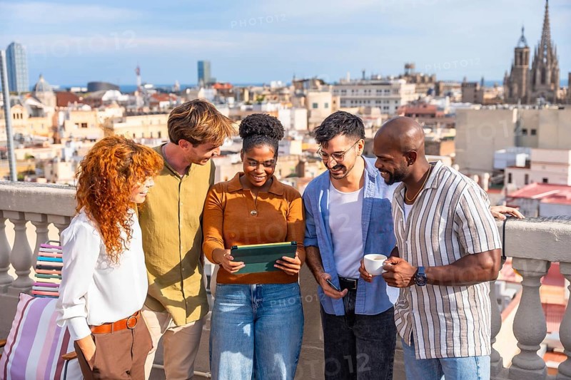 Diverse colleagues networking and smiling on a sunny barcelona rooftop during a casual work break, collaborating and reviewing content on a tablet with city skyline views
