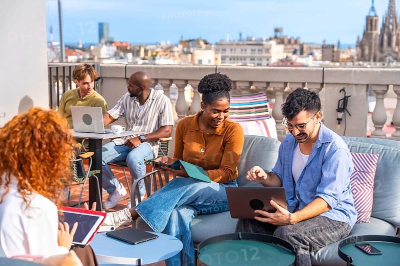 Group of diverse business people meeting and working together casually on a rooftop terrace, discussing ideas and using digital devices in an urban open air office environment