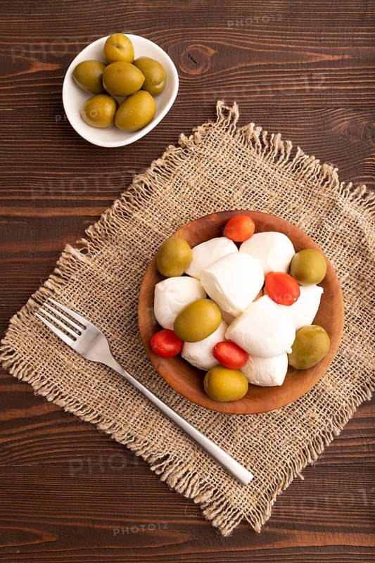 White Mozzarella cheese, with tomatoes and olives in wooden bowl on brown wooden background and linen textile, top view, flat lay, close up