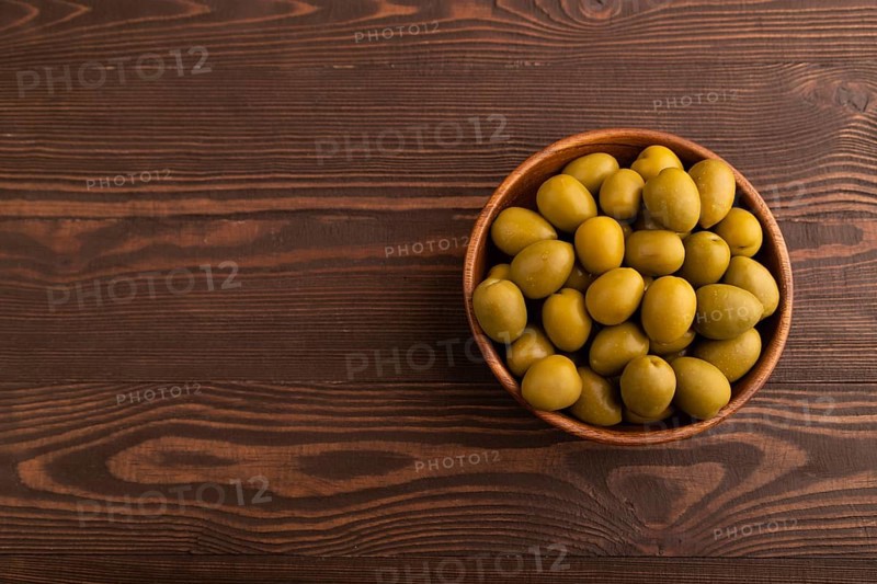 Fresh green olives in wooden bowl on brown wooden background. Top view, flat lay, copy space