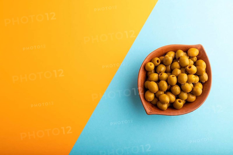 Fresh green olives in ceramic clay bowl on blue and orange pastel paper background. Top view, flat lay, copy space