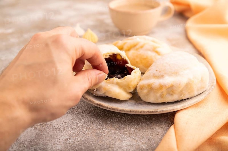 Glazed Pies with blueberry jam on brown concrete background and orange linen textile with hand, cup of coffee, side view, close up, selective focus