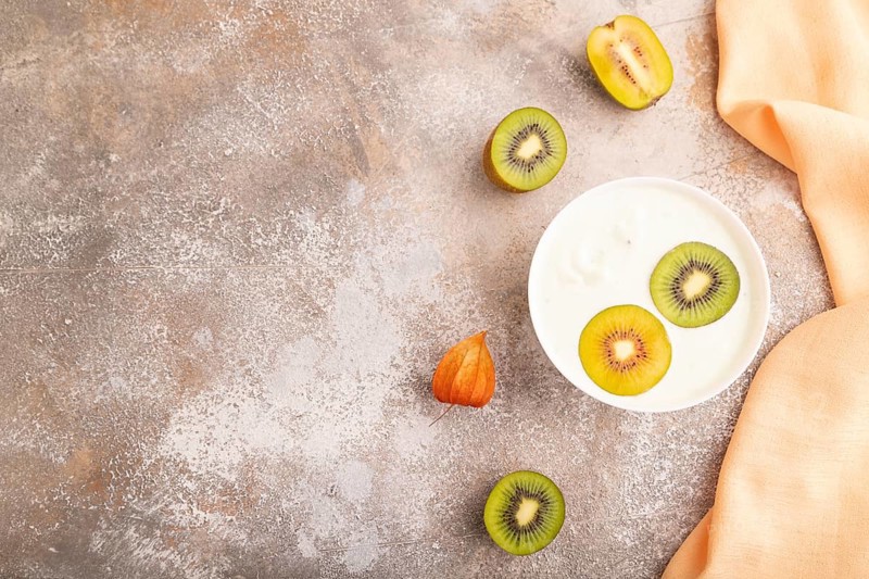 Yogurt with kiwi in white bowl on brown concrete background and orange linen textile, top view, flat lay, copy space, minimalism