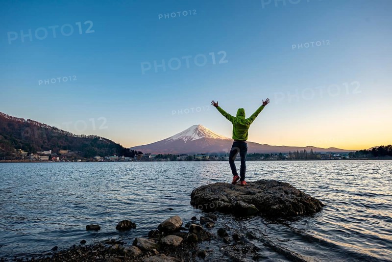 Young man stretching his arms in the air, standing on rocks in Lake Kawaguchi, view of Mount Fuji volcano at sunset, Yamanashi Prefecture, Japan
