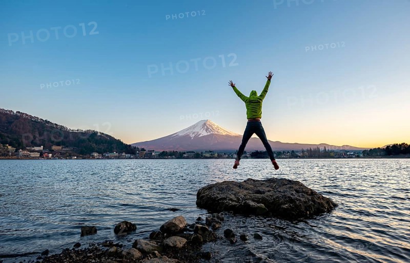 Young man jumping and stretching his arms in the air, standing on rocks in Lake Kawaguchi, view of Mount Fuji volcano at sunset, Yamanashi Prefecture, Japan