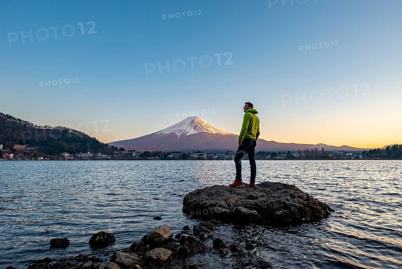 Young man standing on rocks in Lake Kawaguchi, view of Mount Fuji volcano at sunset, Yamanashi Prefecture, Japan