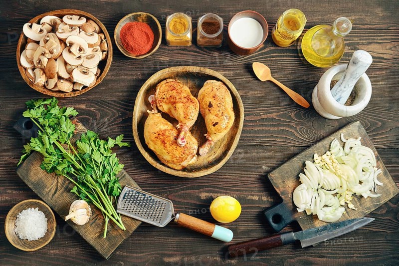 Top view of fried chicken thighs on a wooden plate. Chopped onions, mushrooms, and herbs are placed nearby. Various spices and oil are in jars ready for cooking